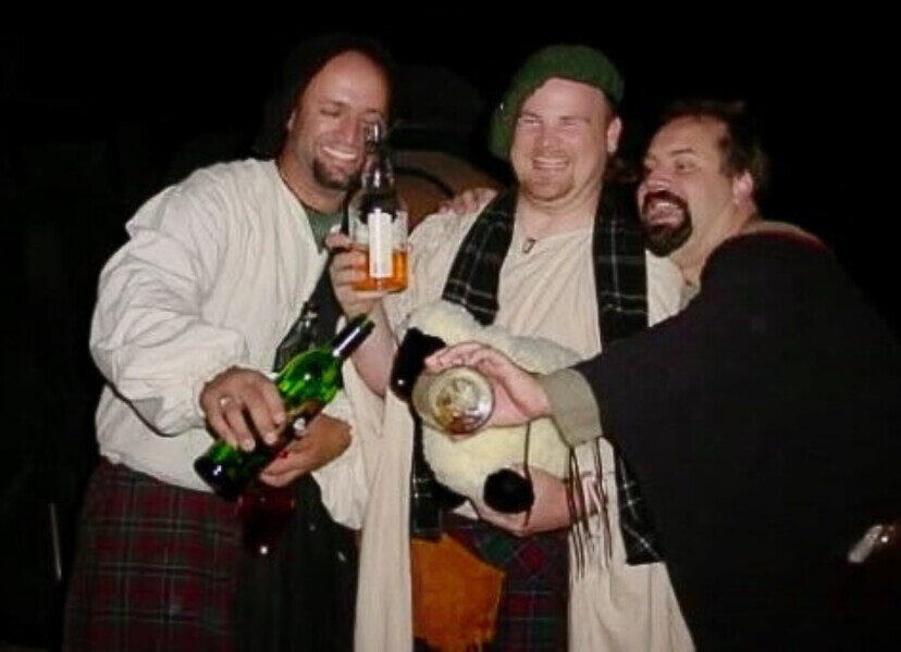 Three laughing men in medieval dress, one holding up a bottle of whisky and one holding a stuffed sheep
