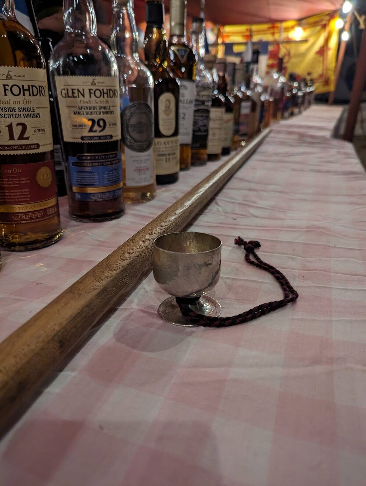 A long table of whisky bottles receding into the distance, a metal tasting cup in the foreground